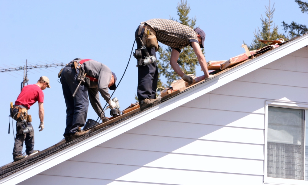 Three construction workers installing a new roof on a house, using nail guns and handling roofing materials on a steep slope.